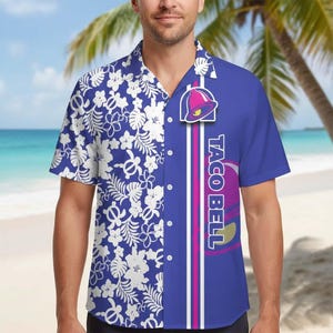 May include: Blue short-sleeve button-down shirt. The left side features a white floral pattern on a blue background. The right side has a Taco Bell logo and text on a blue background with white and pink stripes. The shirt is worn by a person on a beach.