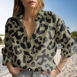 May include: A button-down shirt with a leopard print pattern in black and olive green. The shirt has long sleeves and a collared neckline. The model is wearing the shirt with gray jeans. The background is a beach scene.