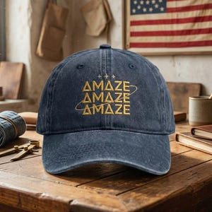 Puede incluir: Gorra de béisbol azul marino con la palabra "AMAZE" bordada en hilo dorado. La gorra tiene una visera curva y un diseño clásico de seis paneles. El fondo incluye una superficie de madera y una bandera americana.