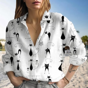Puede incluir: Camisa blanca con botones con un patrón repetido de ilustraciones de gatos negros. La camisa de manga larga tiene cuello y está hecha de una tela ligera. Los diseños de gatos varían en pose y estilo, creando una estética lúdica.