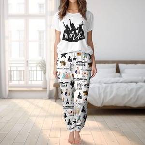 Op de afbeelding: Witte pyjamaset. Het T-shirt heeft het woord "work" en een silhouetontwerp. De pyjamabroek heeft een witte achtergrond met illustraties en tekst, waaronder sterren en het woord "Hamilton".