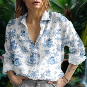 May include: White button-down shirt with a blue tropical print featuring cartoon characters, palm trees, and ocean scenes. The long-sleeved shirt has a collared neckline and is worn open. The print is repeated across the fabric.