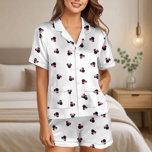 Puede incluir: Conjunto de pijama de satén blanco con un estampado de lazo negro y rosa. La parte superior de manga corta tiene cuello con solapas, dos bolsillos delanteros y botones. Los pantalones cortos a juego completan el conjunto.