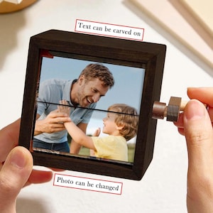 May include: A dark brown wooden photo frame with a rotating mechanism. The frame holds a photograph of a man and a child. The text "Text can be carved on" and "Photo can be changed" are visible on the frame.