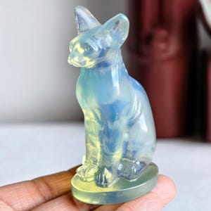 Puede incluir: Una figura de gato translúcida e iridiscente en tonos azules y amarillos. El gato está sentado erguido con las orejas levantadas. La figura está hecha de un material liso y pulido y se asienta sobre una pequeña base redonda.