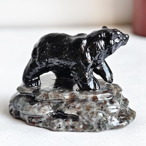Könnte beinhalten: Eine schwarze Bärenfigur aus Stein, die auf einem strukturierten, grau-braunen Sockel steht. Der Bär hat eine glänzende Oberfläche, und der Sockel hat ein natürliches, felsiges Aussehen. Die Figur ist ein Dekorationsartikel.