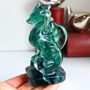 Könnte beinhalten: Eine durchscheinende grüne Fluorite-Drachenskulptur. Der Drache ist mit komplizierten Details geschnitzt, darunter Schuppen und eine fließende Mähne. Die Skulptur steht auf einem dunklen, sechseckigen Sockel. Der Körper des Drachen hat ein wirbelndes Muster aus grünen und klaren Farbtönen.