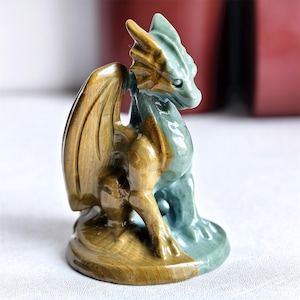 Puede incluir: Pequeña figura de dragón en tonos verdes y dorados. El dragón está sentado con las alas extendidas. La figura está hecha de piedra pulida o mineral, con escamas y detalles.