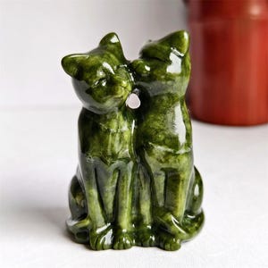 Könnte beinhalten: Eine grüne Steinskulptur von zwei Katzen, von denen eine die andere küsst. Die Katzen sitzen aufrecht, mit einem kleinen rosa Stein zwischen sich. Die Skulptur hat eine polierte, glänzende Oberfläche, vor weißem Hintergrund.