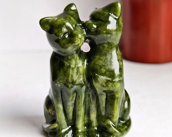 May include: A green stone sculpture of two cats, one kissing the other. The cats are sitting upright, with a small pink stone between them. The sculpture has a polished, glossy finish and is set against a white background.