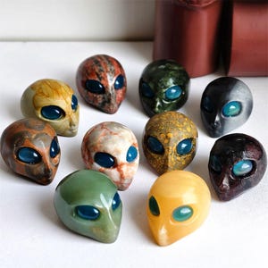 May include: A collection of alien head sculptures in various colors and textures. Each head features large, oval blue eyes and a smooth, rounded form. The colors range from earthy tones to vibrant hues, showcasing unique patterns.
