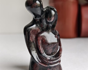 May include: A dark, polished sculpture depicting two figures embracing. The sculpture has a smooth, reflective surface with a deep burgundy and black color. The figures are stylized, with flowing lines and a sense of intimacy.