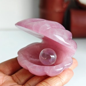 May include: A pink gemstone clam shell with a pearl inside. The clam shell is carved from a translucent pink stone, with a scalloped edge. The pearl is a clear, round sphere with a hint of purple.