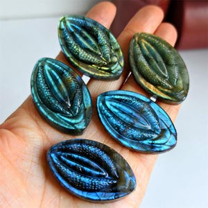 May include: Five oval-shaped labradorite stones with carved leaf-like designs. The stones display iridescent blues, greens, and golds. The stones are held in a hand, showcasing their polished surfaces and intricate details. The overall theme is natural gemstones.