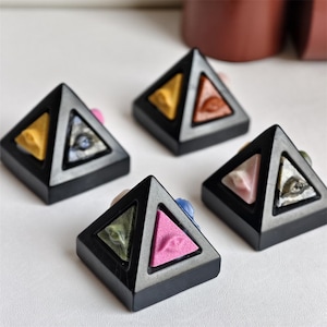Könnte beinhalten: Vier schwarze pyramidenförmige Objekte mit dreieckigen Ausschnitten. Jede Pyramide hat zwei farbige Dreiecke im Inneren: gelb und blau, orange und rosa, grün und rosa sowie weiß und blau. Die Pyramiden befinden sich auf einer weißen Oberfläche.