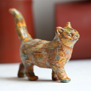 Puede incluir: Una pequeña figura de gato con una superficie texturizada que presenta un patrón jaspeado en colores naranja, marrón y verde azulado apagado. El gato está representado de pie con la cola erguida, mostrando líneas grabadas en todo el cuerpo.
