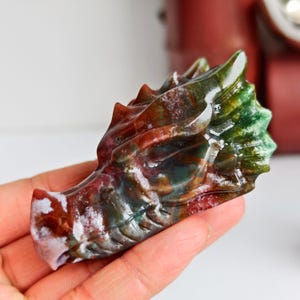 May include: A carved dragon head sculpture in shades of red, green, and white. The gemstone carving features detailed scales and a textured surface. The dragon's head is held in a hand, showcasing the intricate craftsmanship of the stone carving.