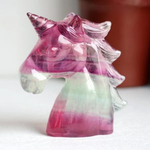 May include: A translucent unicorn figurine with a pink, purple, and green color scheme. The sculpture features a detailed horn, mane, and facial features. The crystal-like material gives it a whimsical and magical appearance, suitable for home decor.