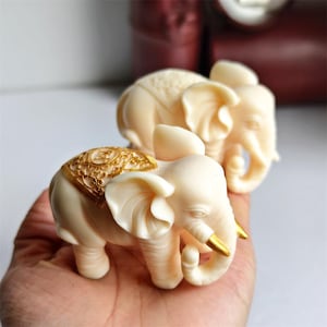 May include: Ivory-colored elephant figurines with gold accents. The elephants have detailed carvings on their backs and trunks. One elephant is in focus, with another slightly blurred in the background. The figurines are held in a hand.