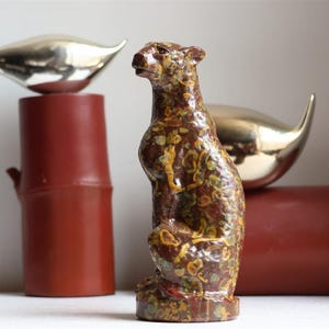 Może przedstawiać: Ceramiczna figurka lamparta z nakrapianą brązowo-żółto-zieloną glazurą, umieszczona przed dwoma cylindrycznymi czerwonymi postumentami, z których każdy zwieńczony jest gładkim, polerowanym, srebrzystym metalowym przedmiotem w kształcie ptaka.