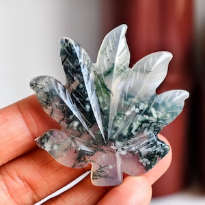 Puede incluir: Una escultura de piedra tallada en forma de hoja. La escultura presenta un diseño detallado con tonos claros, blancos y verdes. Las vetas están intrincadamente talladas, con inclusiones oscuras, casi negras. La piedra tiene un acabado pulido y liso.