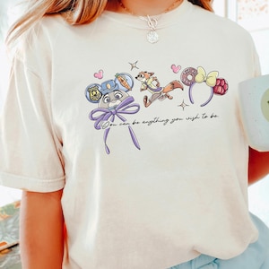 Puede incluir: Camiseta de color marfil con un dise&ntilde;o de dibujos animados caprichoso con personajes y accesorios. El dise&ntilde;o incluye una placa de polic&iacute;a, un zorro y una diadema con forma de rosquilla. Tambi&eacute;n se incluye el texto "You can be anything you wish to be".