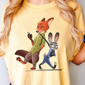 May include: Yellow t-shirt featuring a cartoon illustration of Nick Wilde, a fox, and Judy Hopps, a rabbit, from Zootopia. Nick is holding a phone, and Judy is walking with a leash. The word "BUTTER" is printed at the bottom right.