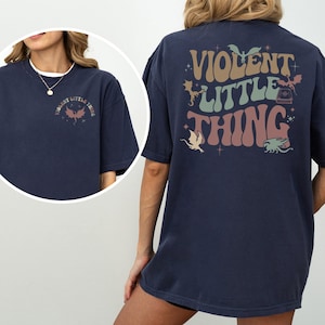 May include: A navy blue t-shirt with the text "VIOLENT LITTLE THING" in a retro font on the back and front. The design includes illustrations of dragons and stars in shades of brown, green, and pink.