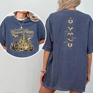 May include: Denim blue t-shirt with a gold graphic of a castle and the text "Riorson House" on the front. The back features a vertical gold design of dragons and clouds. The shirt is a relaxed fit.