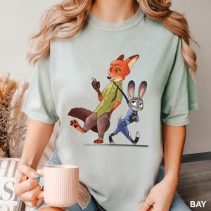 May include: A light green t-shirt featuring a cartoon illustration of Nick Wilde, a fox, and Judy Hopps, a rabbit, from the movie Zootopia. Nick is holding a phone, and Judy is walking beside him. The word "BAY" is printed in white.
