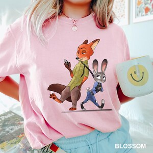 May include: A light pink t-shirt featuring a cartoon illustration of Nick Wilde and Judy Hopps from Zootopia. Nick is holding a phone, and Judy is walking. A white mug with a yellow smiley face is also visible.