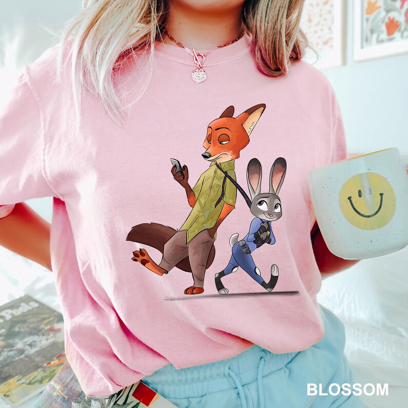 May include: A light pink t-shirt featuring a cartoon illustration of Nick Wilde and Judy Hopps from Zootopia. Nick is holding a phone, and Judy is walking. A white mug with a yellow smiley face is also visible.