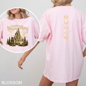 May include: A light pink t-shirt with a graphic of a castle and the text "Riorgon House" on the front. The back of the shirt features a gold design. The shirt is made of a soft material.