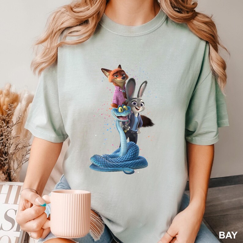 May include: A pale green t-shirt with a cartoon graphic of characters from the film Zootropolis. The design features Nick Wilde the fox, Judy Hopps the rabbit, and a blue snake. The word "BAY" is printed on the bottom right of the t-shirt.