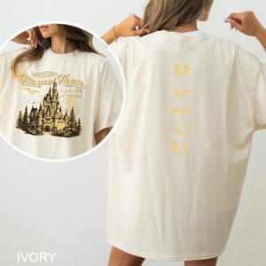 May include: Ivory-colored t-shirt with a graphic of a castle and the text "Riccon House" on the front. The back of the shirt features a vertical design of stylized symbols in a similar color. The shirt is a relaxed fit.