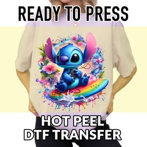 May include: A beige t-shirt featuring a vibrant graphic of a blue cartoon character holding a game controller while surfing on a rainbow-colored surfboard. The image includes the text "READY TO PRESS" and "HOT PEEL DTF TRANSFER".