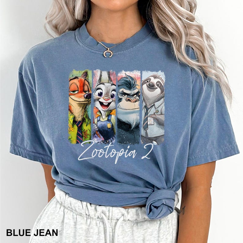 Puede incluir: Camiseta azul vaquero con un gr&aacute;fico colorido de personajes de Zootopia 2. El gr&aacute;fico incluye a Nick Wilde, Judy Hopps, un rinoceronte y Flash el perezoso. El texto "Zootopia 2" est&aacute; escrito en cursiva.