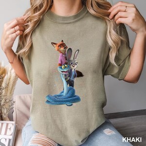May include: Khaki-coloured t-shirt with a vibrant graphic of animated characters: a fox in a pink shirt, a rabbit in a blue jacket, and a blue snake. The word "KHAKI" is printed in white at the bottom right.