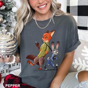 May include: Dark gray t-shirt featuring a cartoon fox and bunny from Zootopia. The fox is wearing a green shirt and brown pants, holding a phone. The bunny is in a blue police uniform. The word "PEPPER" is at the bottom.