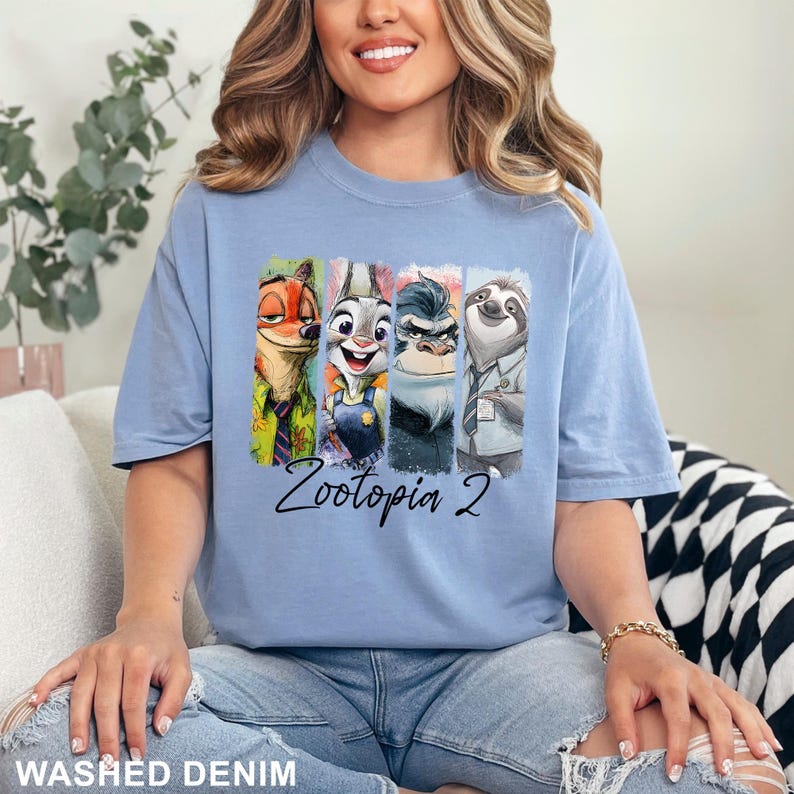 Puede incluir: Una camiseta azul claro con personajes de Zootopia. El dise&ntilde;o muestra a Nick Wilde, Judy Hopps, un gorila y Flash el perezoso en paneles verticales coloridos. Las palabras "Zootopia 2" est&aacute;n escritas en cursiva debajo.