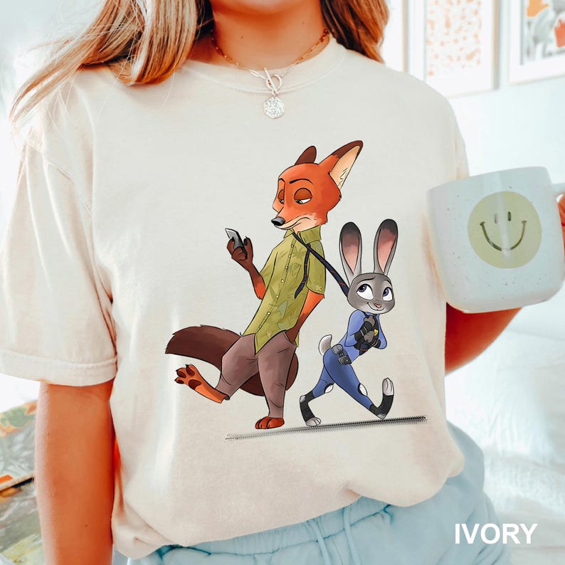 May include: Ivory-colored t-shirt featuring a cartoon illustration of a fox and a rabbit. The fox is holding a phone, and the rabbit is walking on a leash. A white mug with a smiley face is also visible.