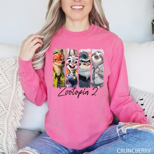 Puede incluir: Una sudadera rosa brillante de manga larga con un gr&aacute;fico colorido de personajes de la pel&iacute;cula animada "Zootr&oacute;polis". El gr&aacute;fico incluye a Nick Wilde, Judy Hopps, el jefe Bogo y Flash, con el texto "Zootopia 2" debajo.