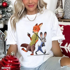 May include: White t-shirt featuring a cartoon illustration of Nick Wilde the fox and Judy Hopps the rabbit from Zootopia. Nick is holding a phone, and Judy is pulling him along. The word "WHITE" is printed at the bottom.