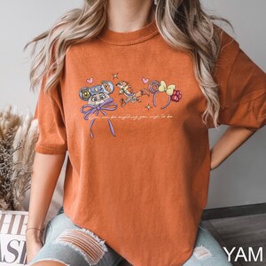 Puede incluir: Camiseta naranja con un dise&ntilde;o de dibujos animados de un rat&oacute;n, un zorro y un lazo, con el texto "You can be anything you wish to be." El dise&ntilde;o es de estilo caprichoso.