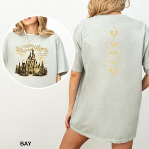 May include: A light green t-shirt with a graphic of a castle and the text "Biscay Home" on the front. The back of the shirt features a vertical design with various symbols in a gold color. The shirt is a relaxed fit.