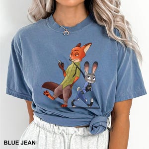 May include: Blue jean colored t-shirt featuring a cartoon illustration of Nick Wilde and Judy Hopps from Zootopia. Nick is a fox wearing a green shirt and Judy is a rabbit in a blue uniform. The text "BLUE JEAN" is at the bottom.