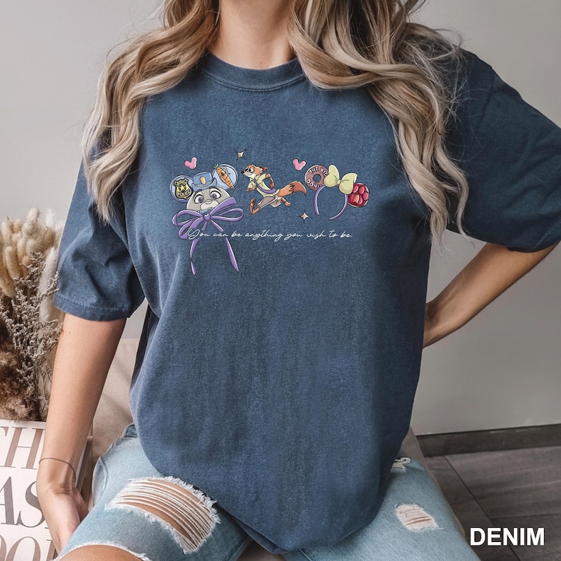 Puede incluir: Camiseta azul denim con un gr&aacute;fico caprichoso de diademas de personajes de dibujos animados y el texto "You can be anything you wish to be." El dise&ntilde;o incluye una diadema de orejas de rat&oacute;n, una ardilla y una diadema de frambuesa.