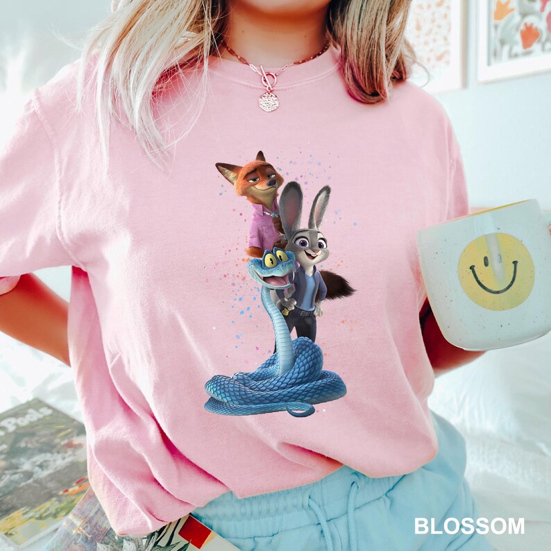 May include: A pale pink t-shirt with a vibrant graphic of characters from the animated film Zootropolis. The design includes a fox, a rabbit, and a snake. A white mug with a yellow smiley face is held in the hand.