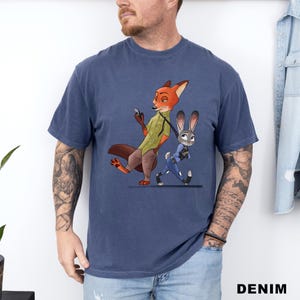 May include: A navy blue t-shirt featuring a cartoon illustration of Nick Wilde and Judy Hopps from Zootopia. Nick, an orange fox, is holding a phone, while Judy, a gray rabbit, walks beside him. The word "DENIM" is printed at the bottom.