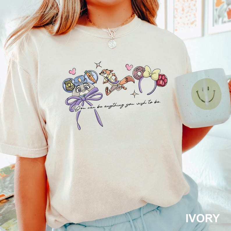 Puede incluir: Camiseta de color marfil con un dise&ntilde;o de dibujos animados caprichoso con personajes y accesorios. El dise&ntilde;o incluye una placa de polic&iacute;a, un zorro y una diadema con forma de rosquilla. Tambi&eacute;n se incluye el texto "You can be anything you wish to be".